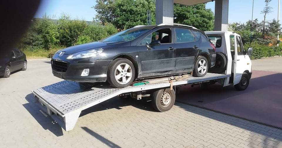 Czarny Peugeot kombi na platformie lawety z otwartymi drzwiami, na stacji benzynowej.