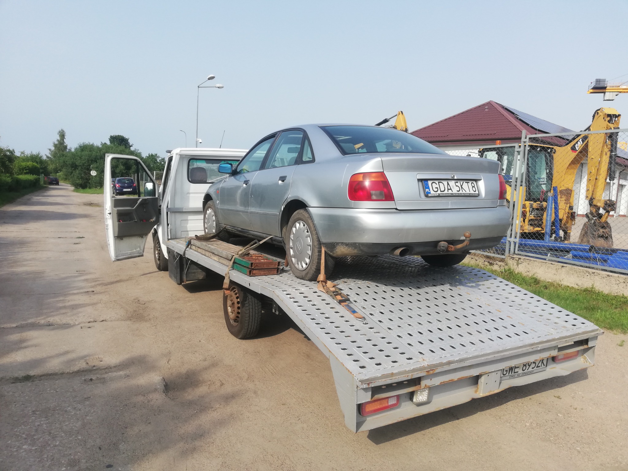 Srebrny samochód osobowy marki Audi A4 na platformie lawety, z otwartymi drzwiami kabiny i widocznymi tablicami rejestracyjnymi, w tle plac budowy z koparką.