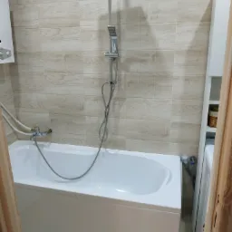 Nowoczesna, wykafelkowana łazienka z białą wanną, baterią wannową, deszczownicą i grzejnikiem gazowym, wykończona płytkami imitującymi drewno.