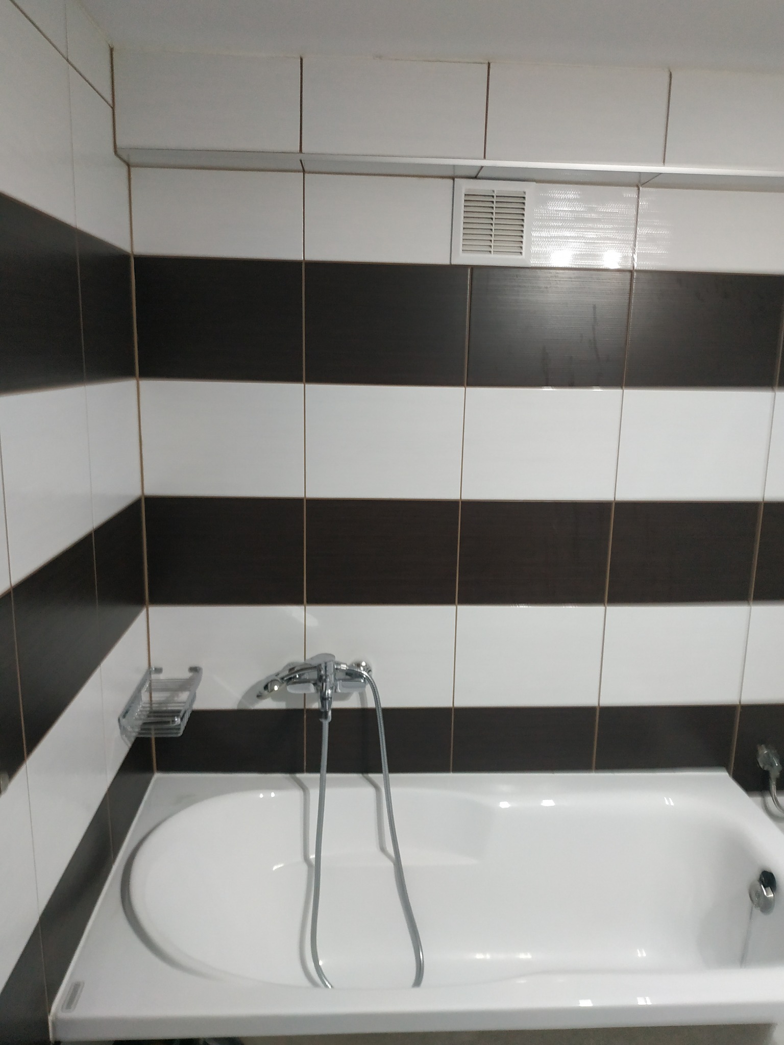 Biała wanna z chromowaną baterią i półką na mydło, otoczona ścianą wyłożoną białymi i brązowymi płytkami w poziome pasy. Widoczna kratka wentylacyjna pod sufitem.