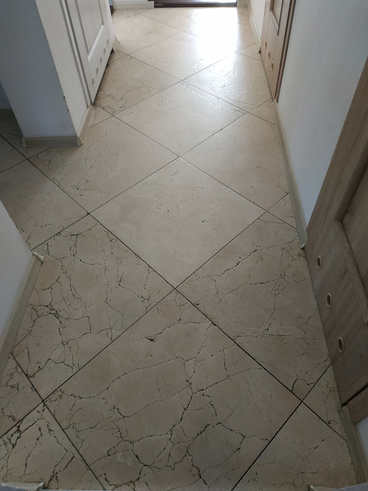 Beżowa podłoga wyłożona marmurkowymi płytkami ułożonymi w karo, widok z góry na fragment przedpokoju z drzwiami po obu stronach.