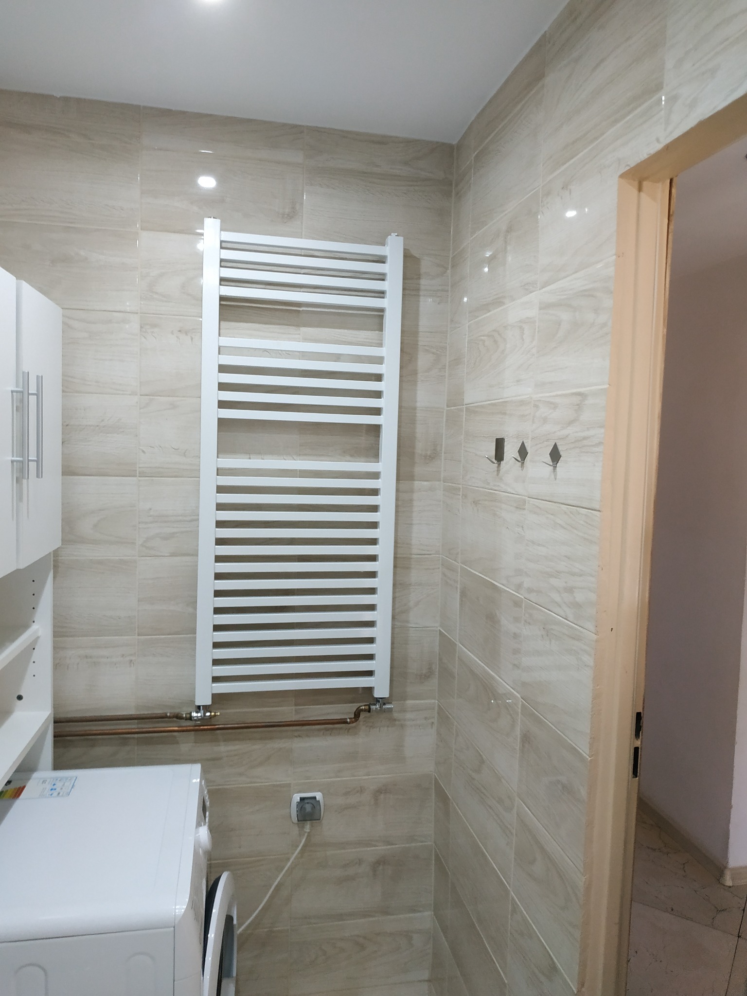 Wykończona łazienka z beżowymi płytkami imitującymi drewno, białym grzejnikiem drabinkowym, pralką i szafką, widoczne również trzy wieszaki na ścianie.