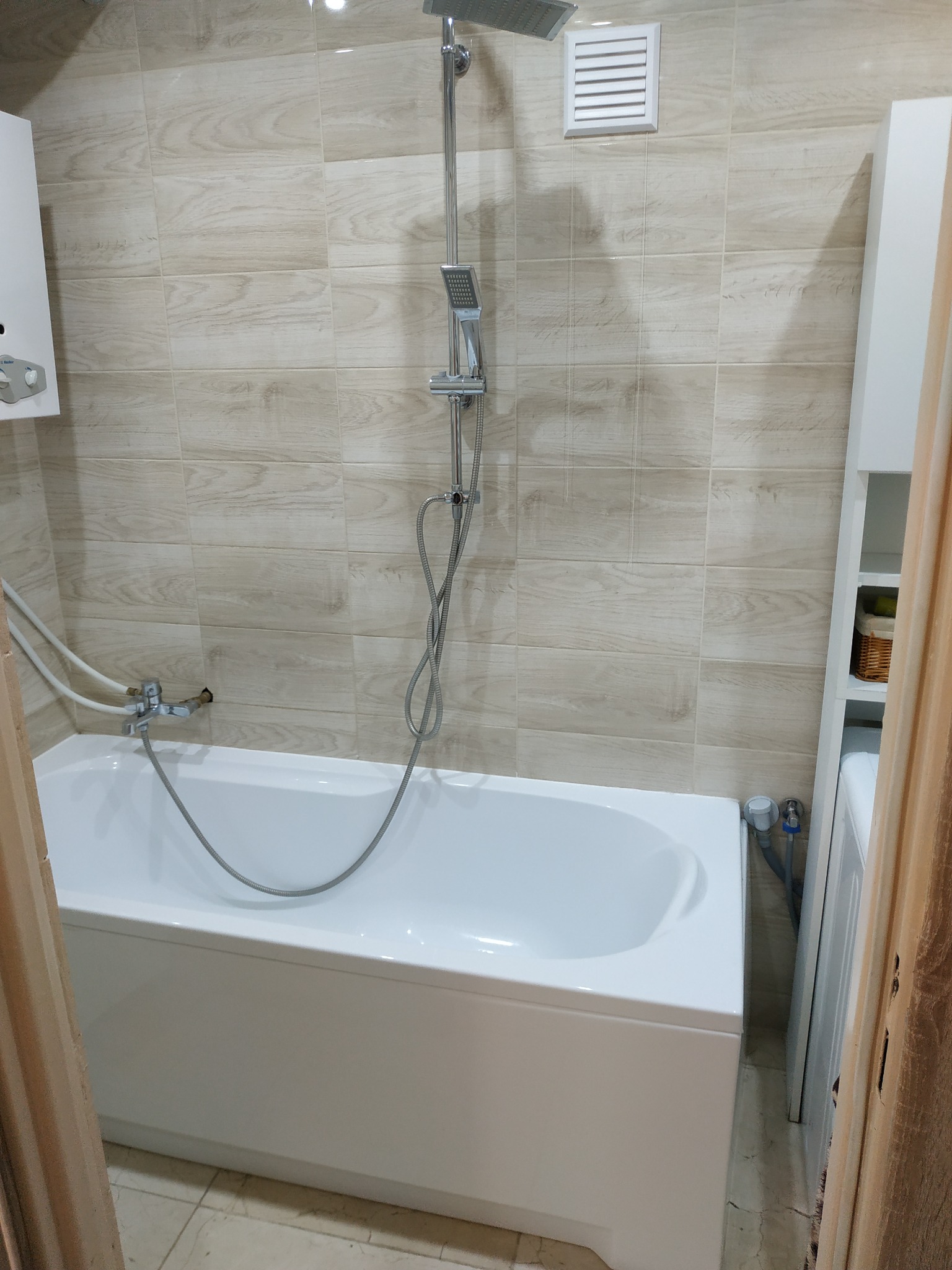 Nowoczesna, wykafelkowana łazienka z białą wanną, baterią wannową, deszczownicą i grzejnikiem gazowym, wykończona płytkami imitującymi drewno.