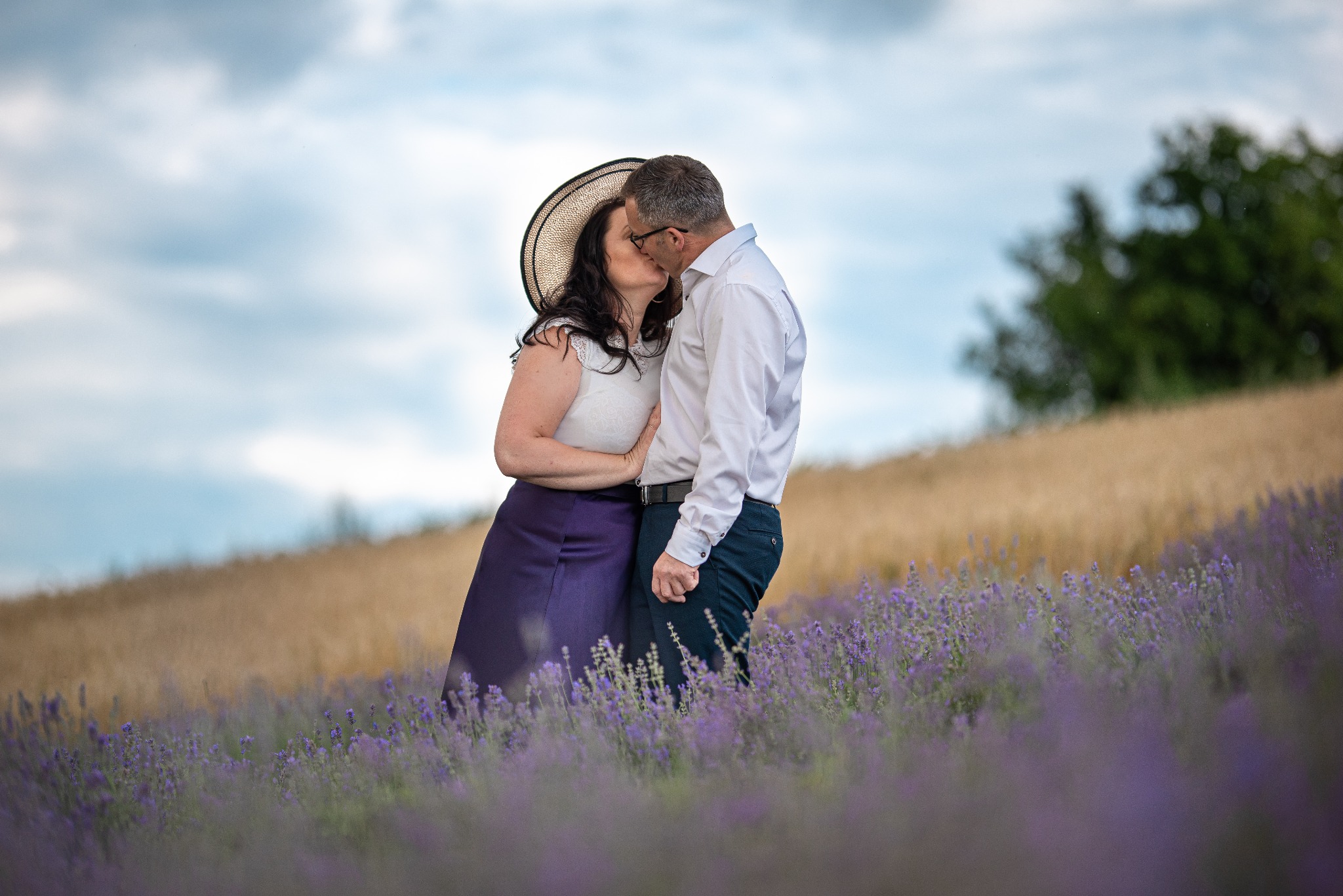Zakochana para całuje się na fioletowym polu lawendy, kobieta w kapeluszu zakrywa część twarzy.