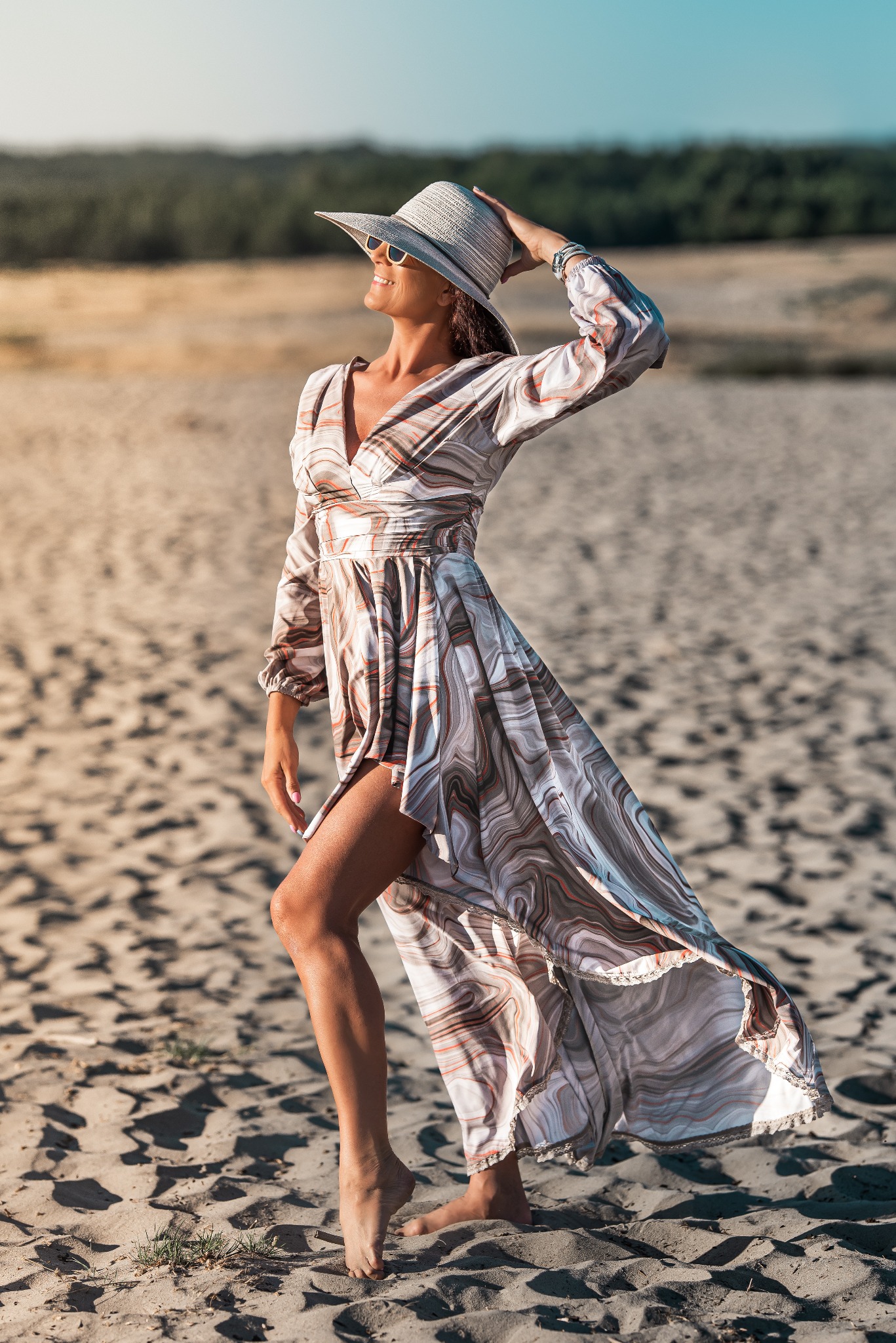 Kobieta w długiej, wzorzystej sukience i kapeluszu pozuje na piaszczystej plaży, trzymając kapelusz i uśmiechając się w okularach przeciwsłonecznych.