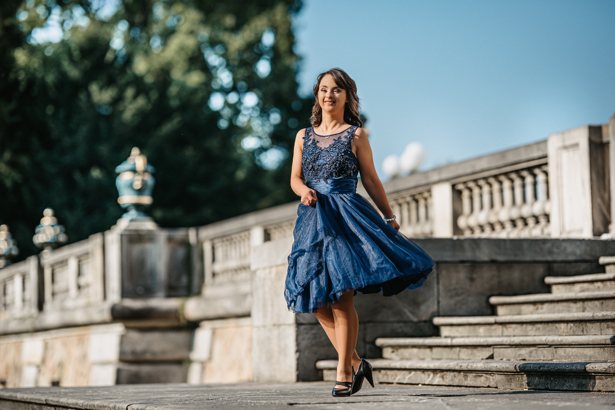 Uśmiechnięta kobieta w granatowej, zwiewnej sukience wiruje na tle kamiennych schodów i balustrady w parku.