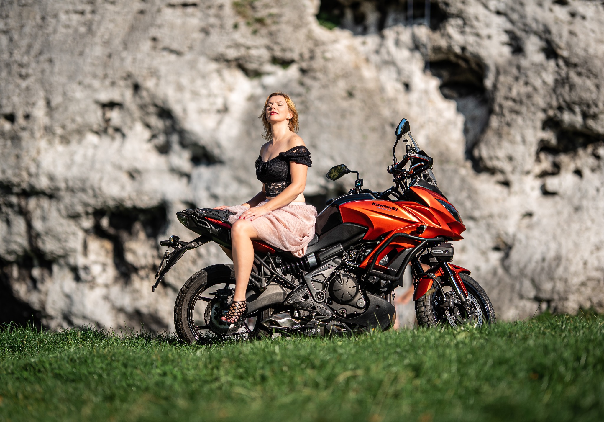 Kobieta w czarnym topie i różowej spódnicy siedzi na pomarańczowym motocyklu Kawasaki na tle skalnej ściany i zielonej trawy.