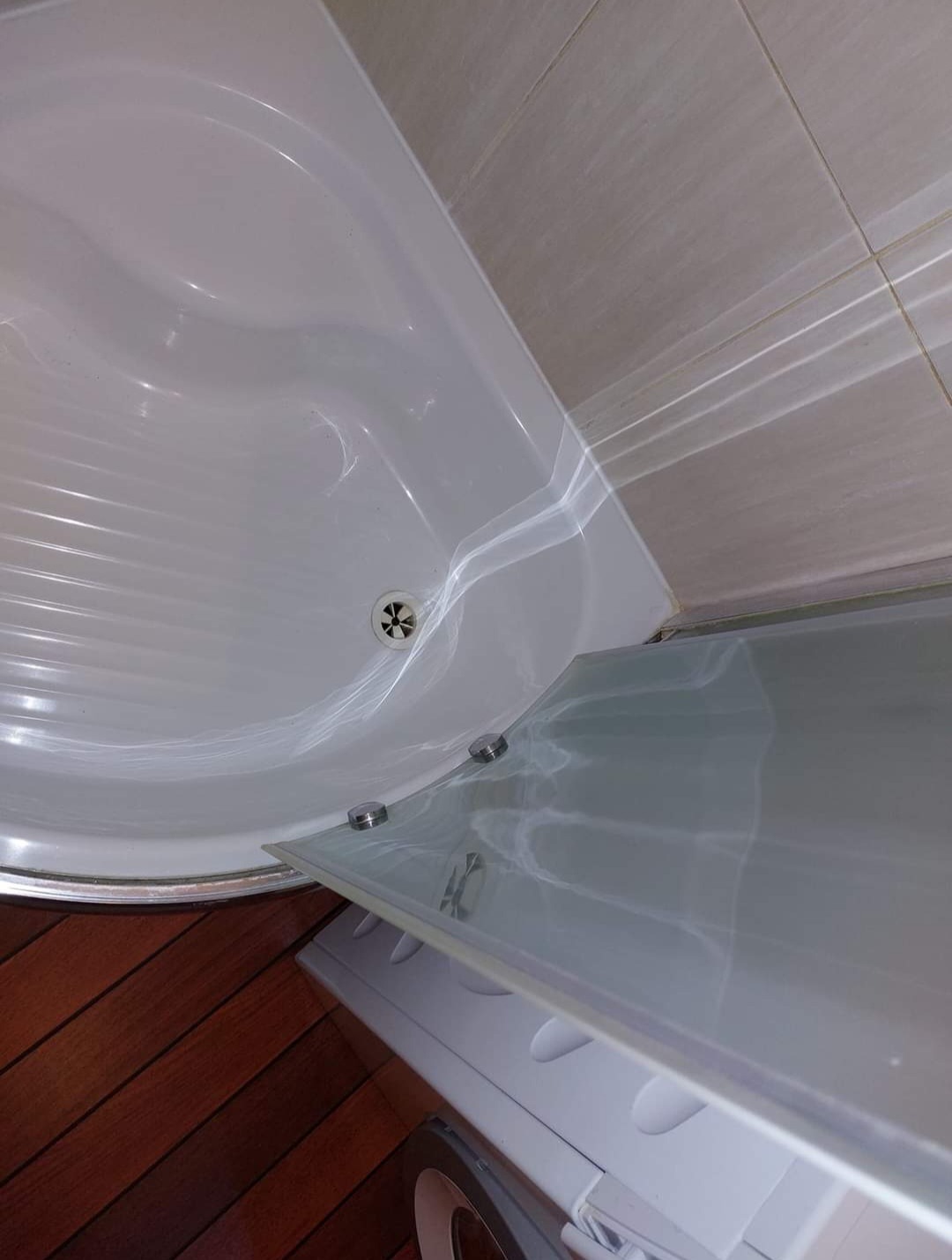 Biały brodzik kabiny prysznicowej z odpływem, częściowo zasłonięty szklanym panelem, widoczne jasne kafelki na ścianie i fragment pralki z boku.