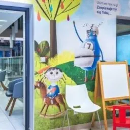 Wnętrze poczekalni z kolorowym muralem przedstawiającym postacie z bajek, krzesła dla dzieci i dorosłych, tablica do rysowania, widok na korytarz z roślinami.