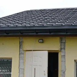 Fragment domu z nowym, grafitowym dachem z dachówki ceramicznej i orynnowaniem, widoczne niedokończone obramowania okien i drzwi.