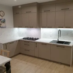 Nowoczesna kuchnia z beżowymi szafkami, blatem imitującym marmur i czarnymi uchwytami, oświetlona listwami LED pod szafkami wiszącymi.