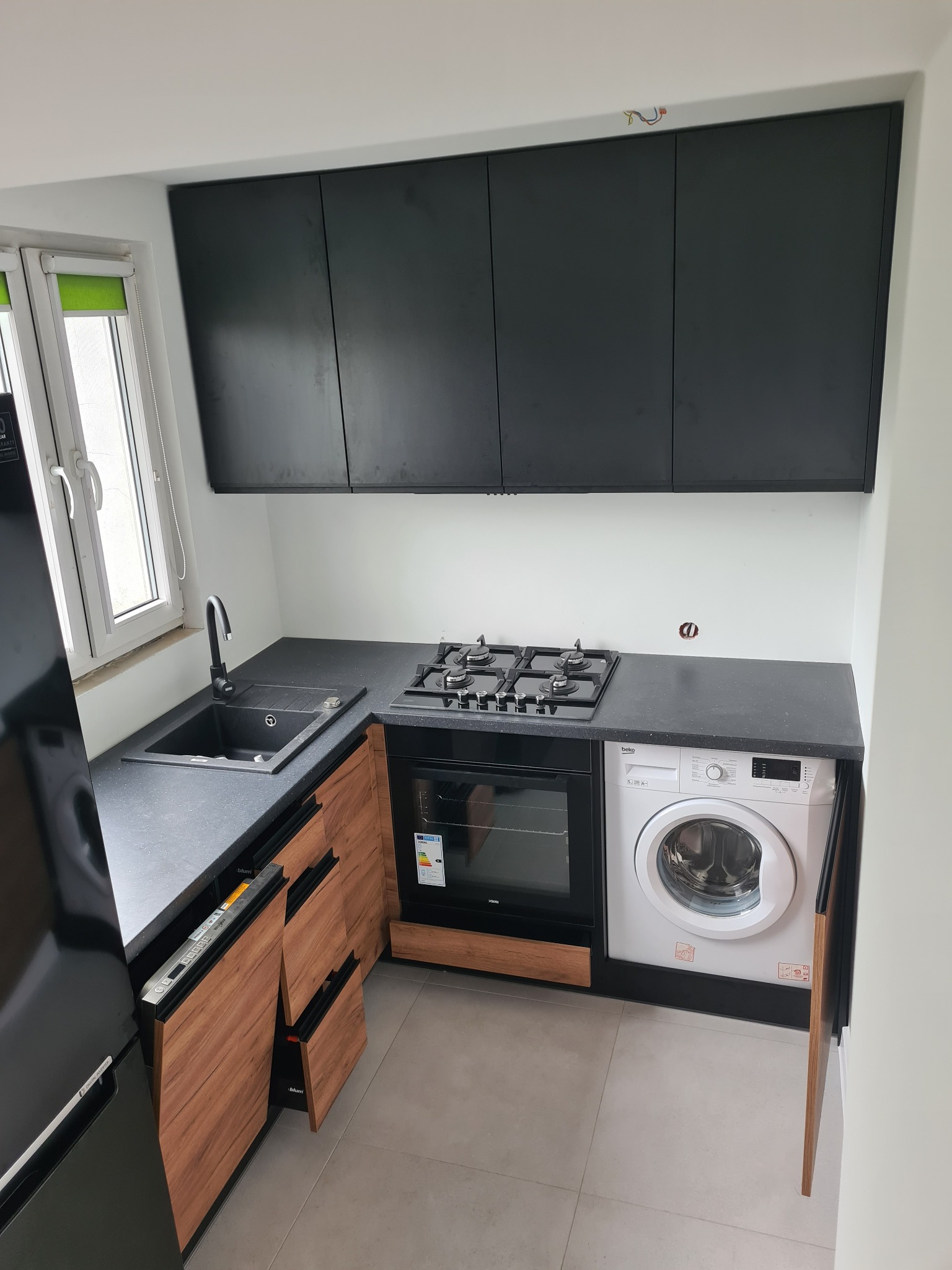 Nowoczesna kuchnia z czarnymi górnymi szafkami, blatem z ciemnego kamienia, drewnianymi frontami dolnych szafek, zintegrowaną pralką i piekarnikiem gazowym.