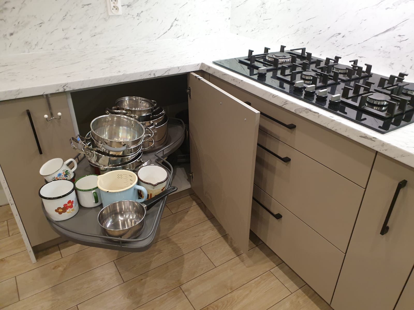 Narożna szafka kuchenna z wysuwaną półką obrotową, wypełniona garnkami i emaliowanymi kubkami, widoczna płyta gazowa na blacie.