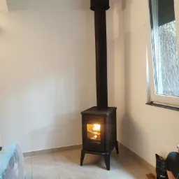 Czarny piec wolnostojący z palącym się drewnem wewnątrz, z widoczną rurą odprowadzającą dym, ustawiony w rogu pomieszczenia z białymi ścianami i oknem.