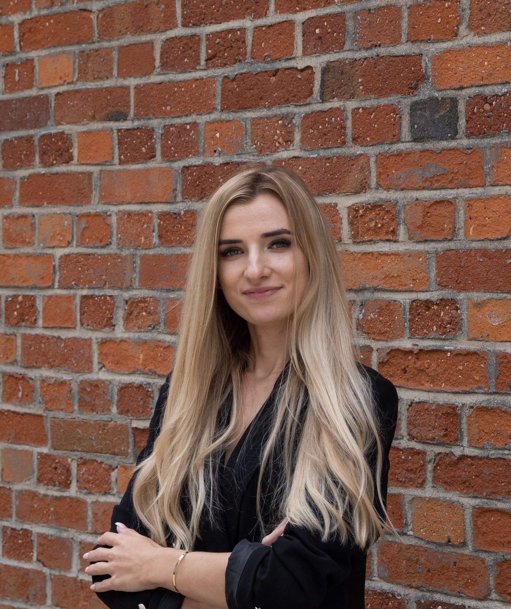 Portret uśmiechniętej blondynki w czarnej marynarce, stojącej na tle ściany z czerwonej cegły. Ręce skrzyżowane, na nadgarstku złota bransoletka.