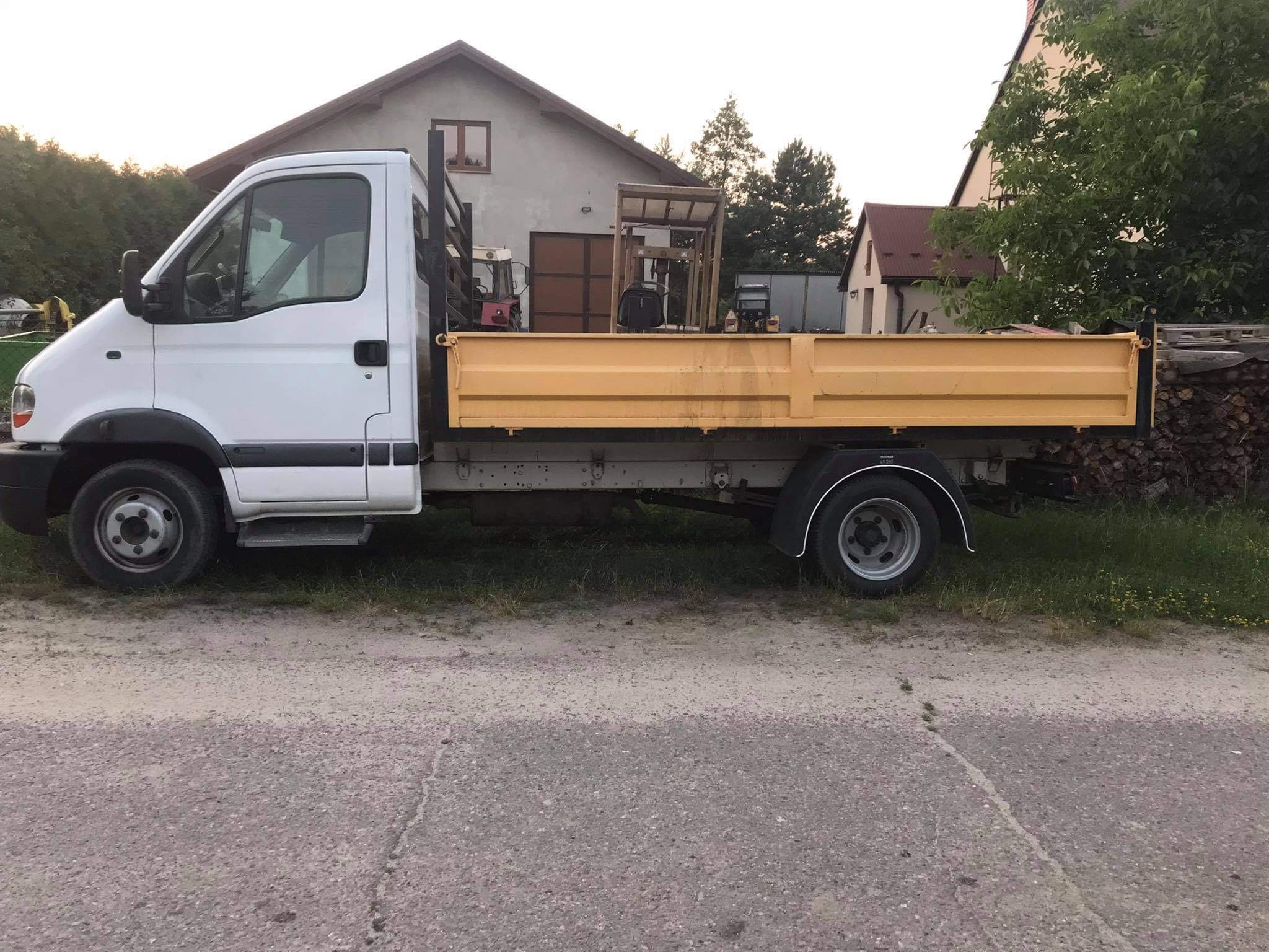 Biały bus z żółtą skrzynią ładunkową zaparkowany na szarej nawierzchni, w tle domy i drzewa.