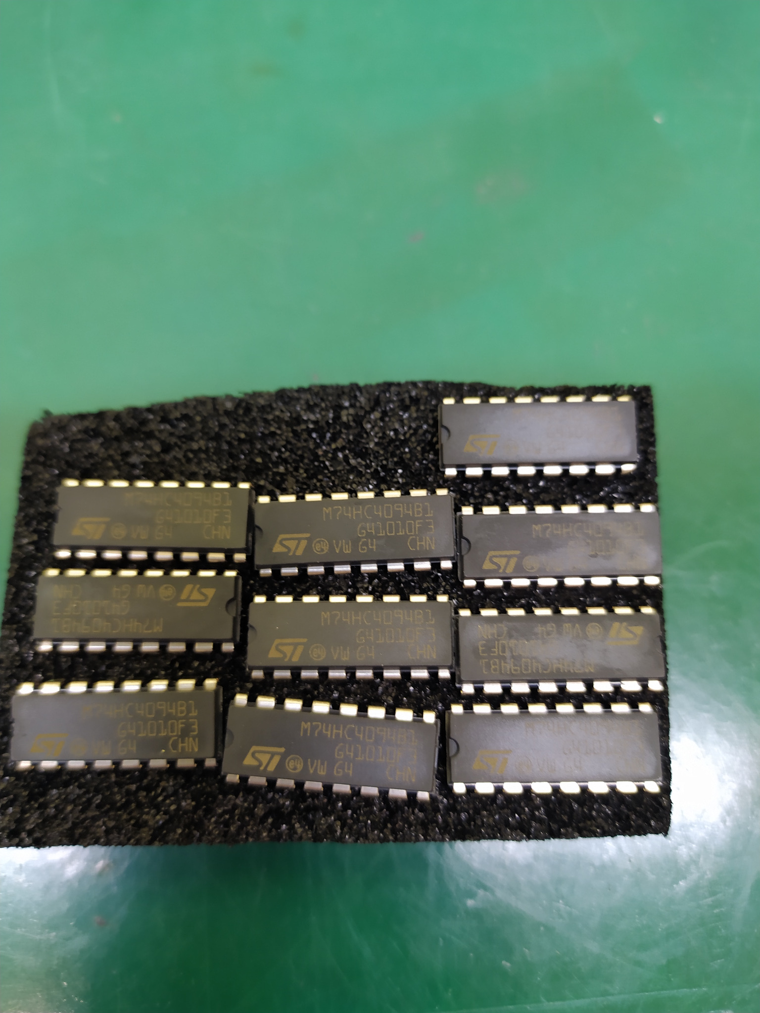 Układ scalony M74HC4094B1 i inne układy scalone STMicroelectronics ułożone na czarnej piance na zielonym tle.