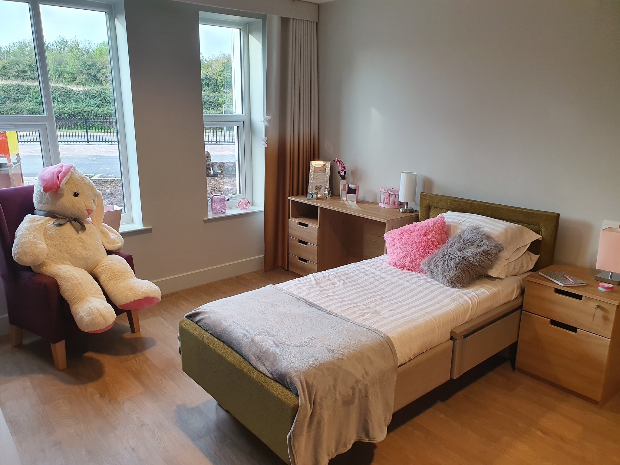 Jasny pokój z pojedynczym łóżkiem, biurkiem i dużym pluszowym misiem siedzącym na krześle, widok z okna na zieleń.