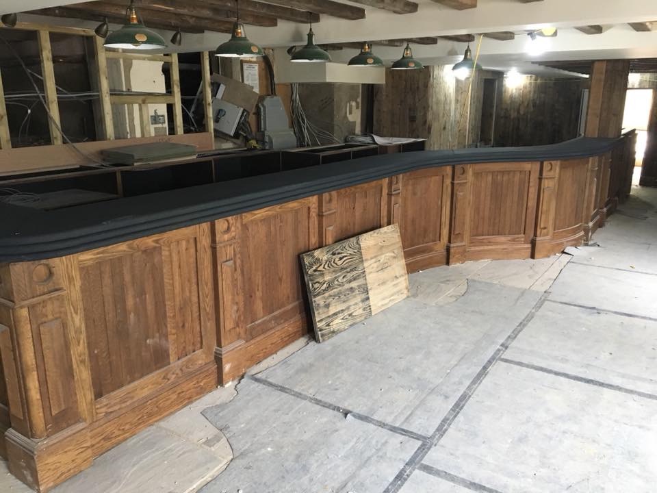 Drewniany bar w trakcie renowacji, widoczne odsłonięte belki stropowe, wiszące lampy i fragment podłogi z widocznymi śladami prac wykończeniowych.