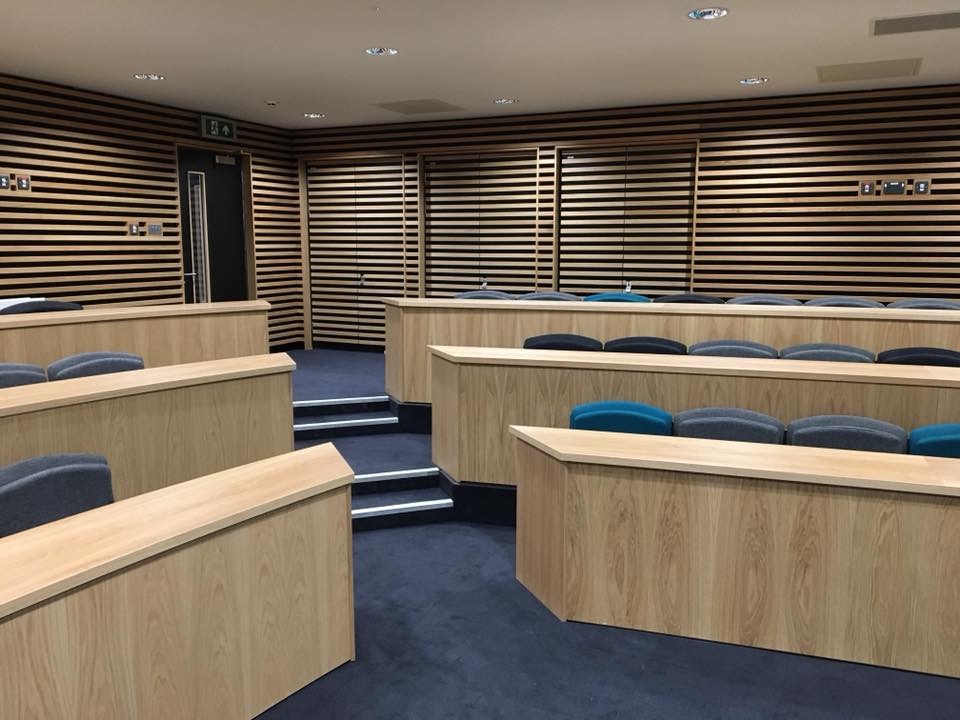 Aula wykładowa z drewnianymi panelami ściennymi i rzędami drewnianych ławek z szarymi i turkusowymi siedziskami, oświetlona światłem punktowym.