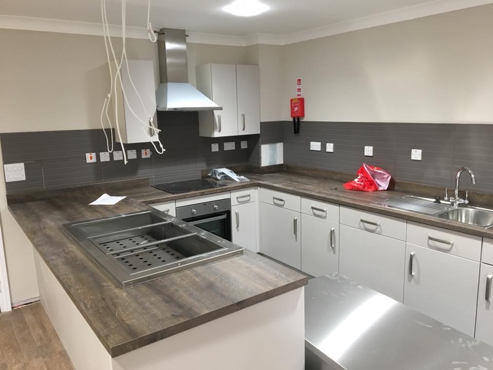 Nowoczesna kuchnia przemysłowa z białymi szafkami, ciemnoszarym backsplashiem i blatem imitującym drewno, wyposażona w stalowy okap, zlewozmywak i pojemnik na żywność w blacie.