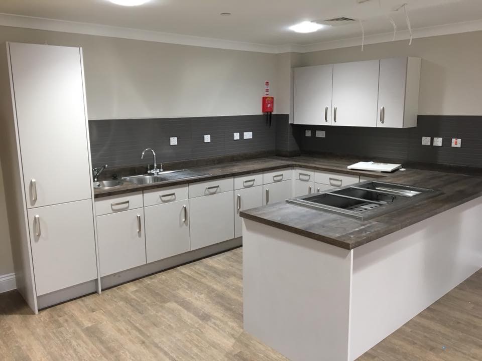 Jasna kuchnia z wyspą i wbudowanym urządzeniem grzewczym, szare płytki nad blatem, podłoga imitująca drewno.