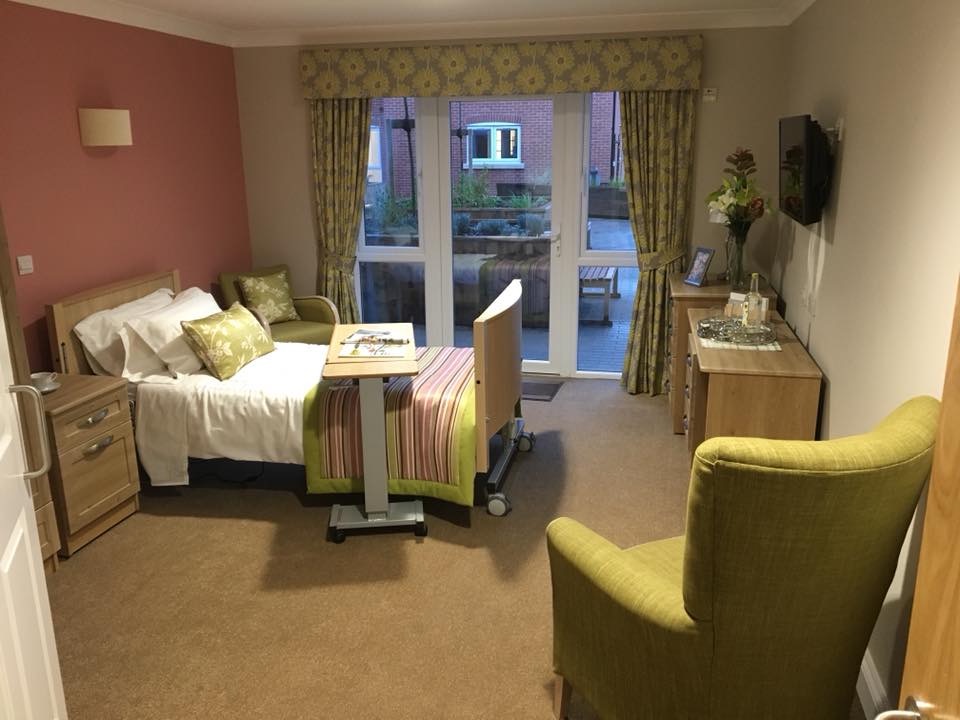 Jasny pokój w domu opieki z łóżkiem, stolikiem na kółkach, komodą z kwiatami i telewizorem, widok na ogród przez przeszklone drzwi balkonowe z zasłonami w kwiaty.