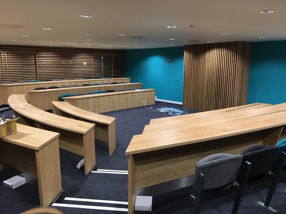 Sala konferencyjna lub audytoryjna z rzędami drewnianych stołów i krzeseł, niebieska ściana w tle i drewniane panele dekoracyjne.