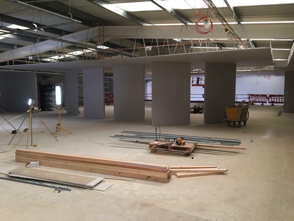 Wnętrze biurowe w trakcie budowy: widoczne surowe ściany, podwieszony sufit w trakcie montażu, materiały budowlane i oświetlenie robocze na podłodze.