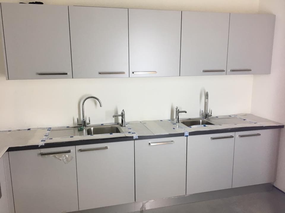 Szara zabudowa kuchenna z dwoma zlewozmywakami i bateriami, blaty zabezpieczone folią ochronną, widoczne metalowe uchwyty.