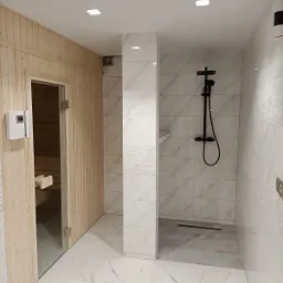 Nowoczesna łazienka z sauną, wykończona białymi płytkami z marmurkowym wzorem, czarna armatura prysznicowa, drewniane wykończenie sauny z szklanymi drzwiami.