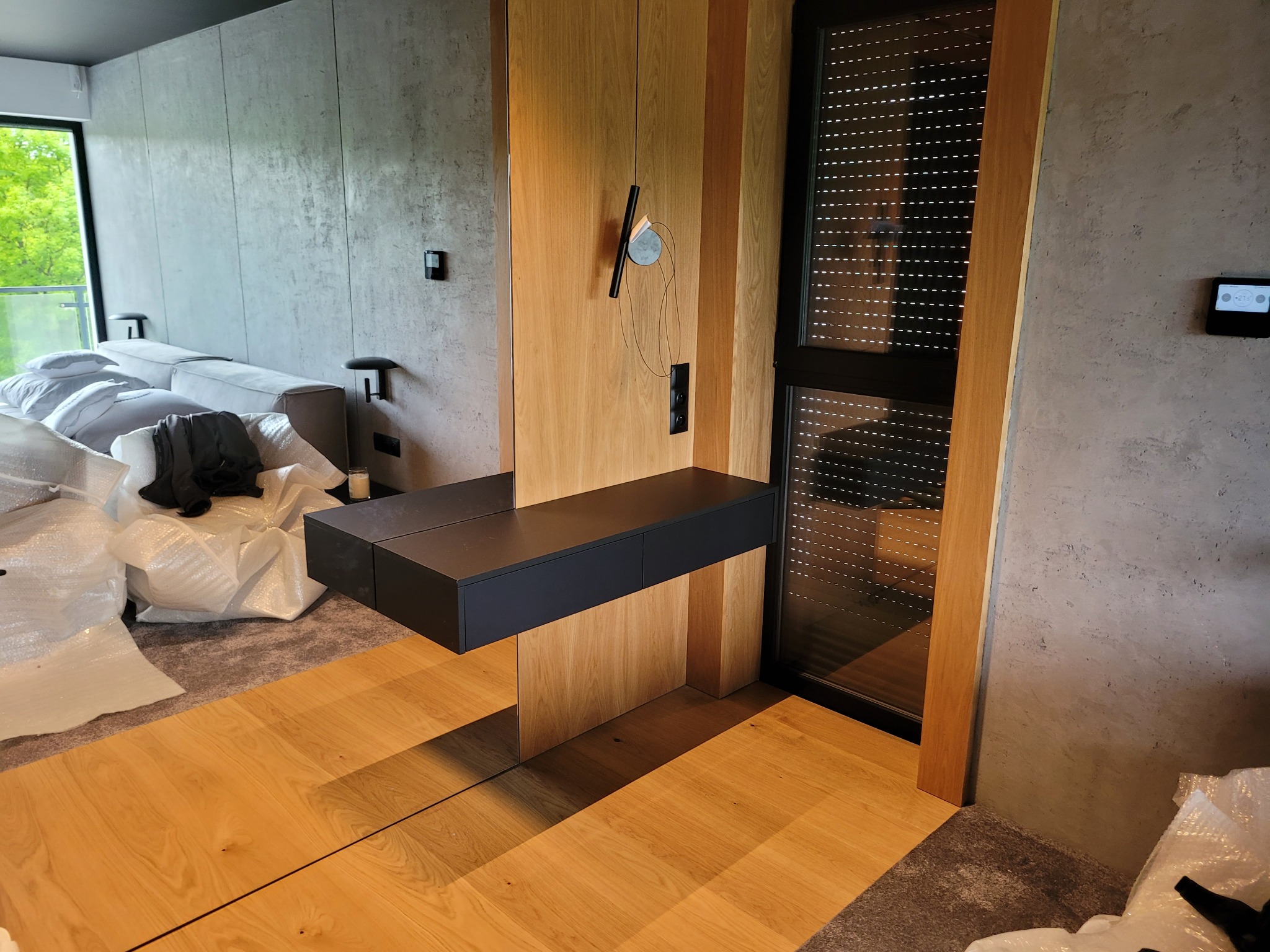 Nowoczesna aranżacja wnętrza z drewnianą zabudową, czarnym blatem i roletami w oknie. Elementy wykończenia w surowym, betonowym stylu. Widoczne materiały zabezpieczające.
