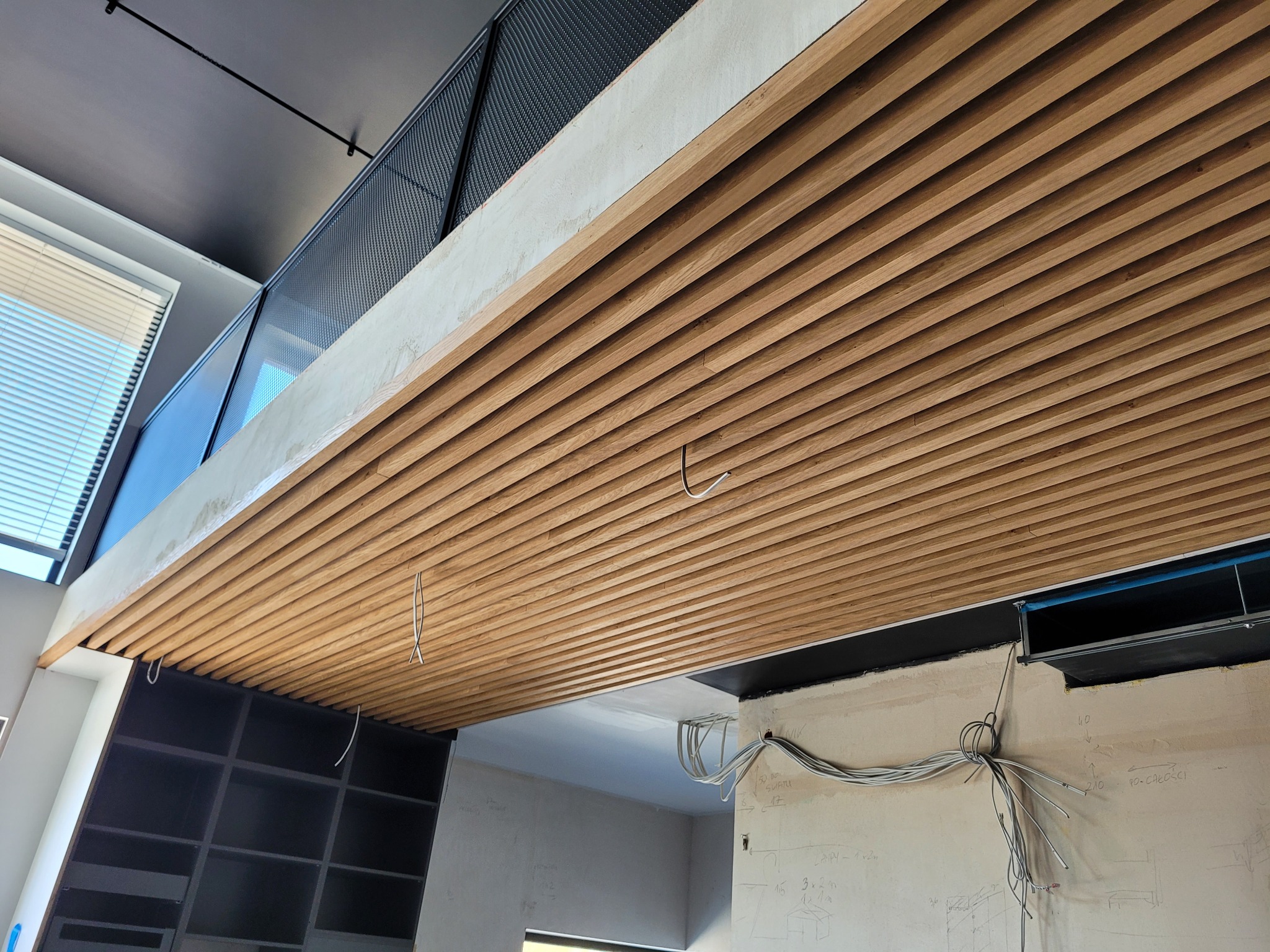 Drewniany sufit lamelowy z widocznymi przewodami, surowa ściana z instalacją elektryczną i notatkami. Widok z dołu, perspektywa wnętrza w trakcie wykończenia.
