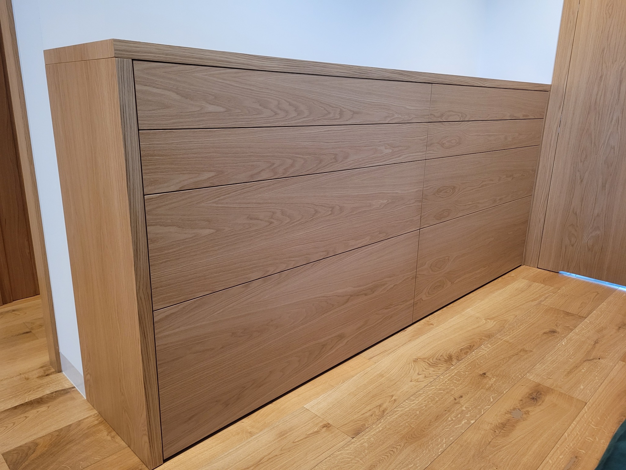 Drewniana zabudowa meblowa z sześcioma szufladami, zintegrowana ze ścianą i podłogą z parkietu. Minimalistyczny design, jasne drewno, subtelne oświetlenie pod szafką.