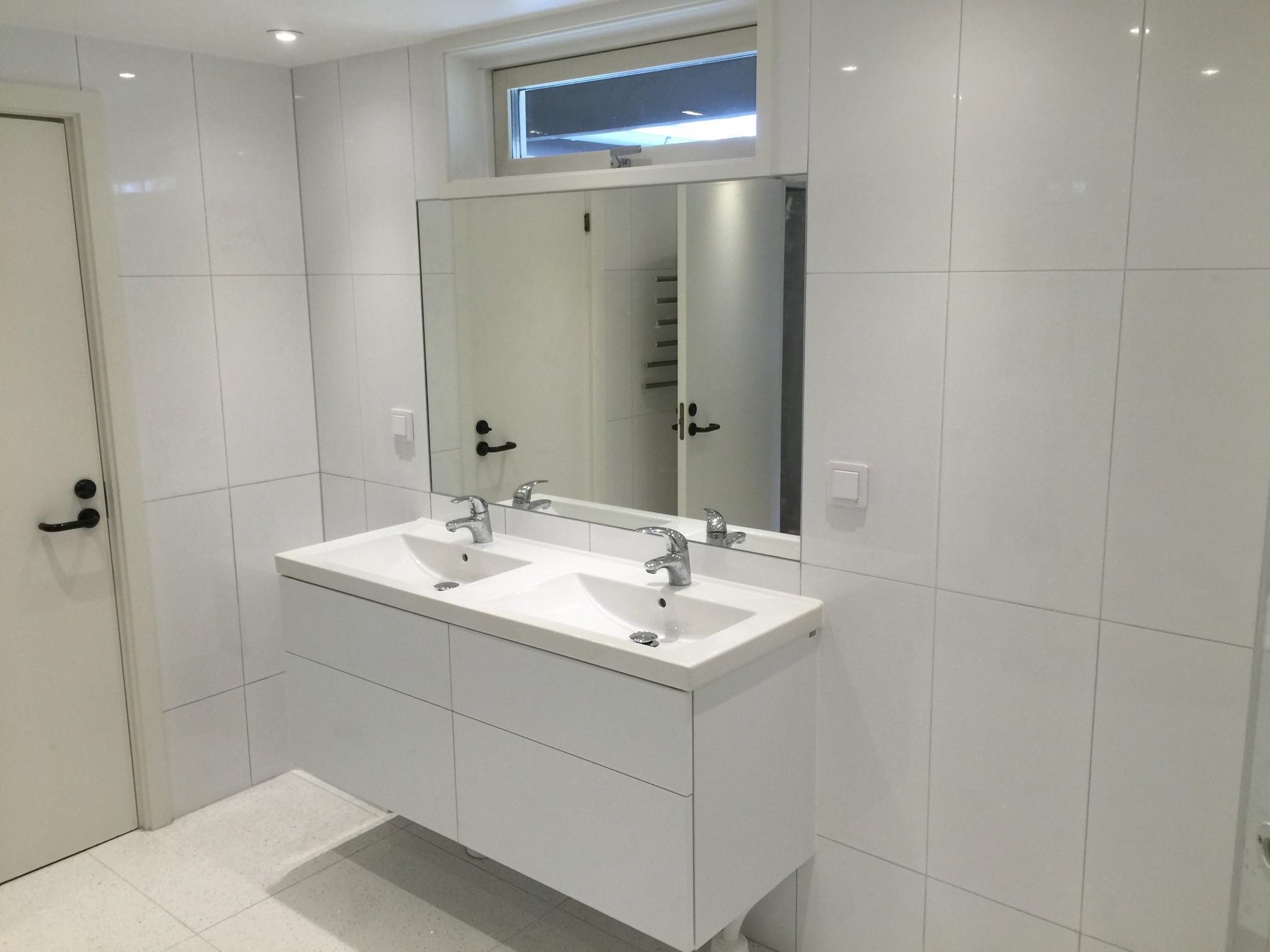 Jasna łazienka w stylu skandynawskim z podwójną umywalką na szafce i białymi płytkami na ścianach oraz podłodze, widoczne odbicie drzwi w lustrze.