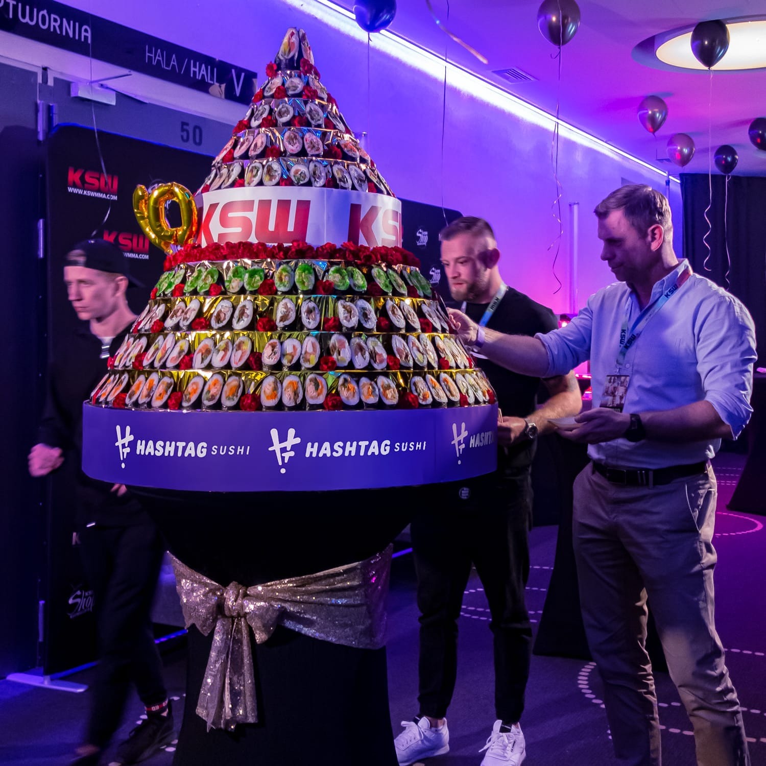 Wielopoziomowy tort z sushi udekorowany różami, ustawiony na stole z błyszczącą kokardą, w tle mężczyźni w sali eventowej z balonami i fioletowym oświetleniem, widoczne logo #HashtagSushi.