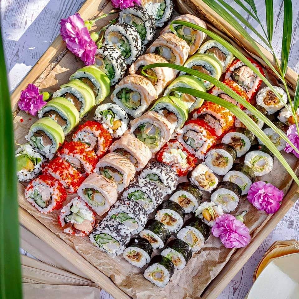 Różnorodny zestaw sushi na drewnianej tacy, ozdobiony kwiatami i zielonymi liśćmi, z widoczną fakturą papieru pod spodem.