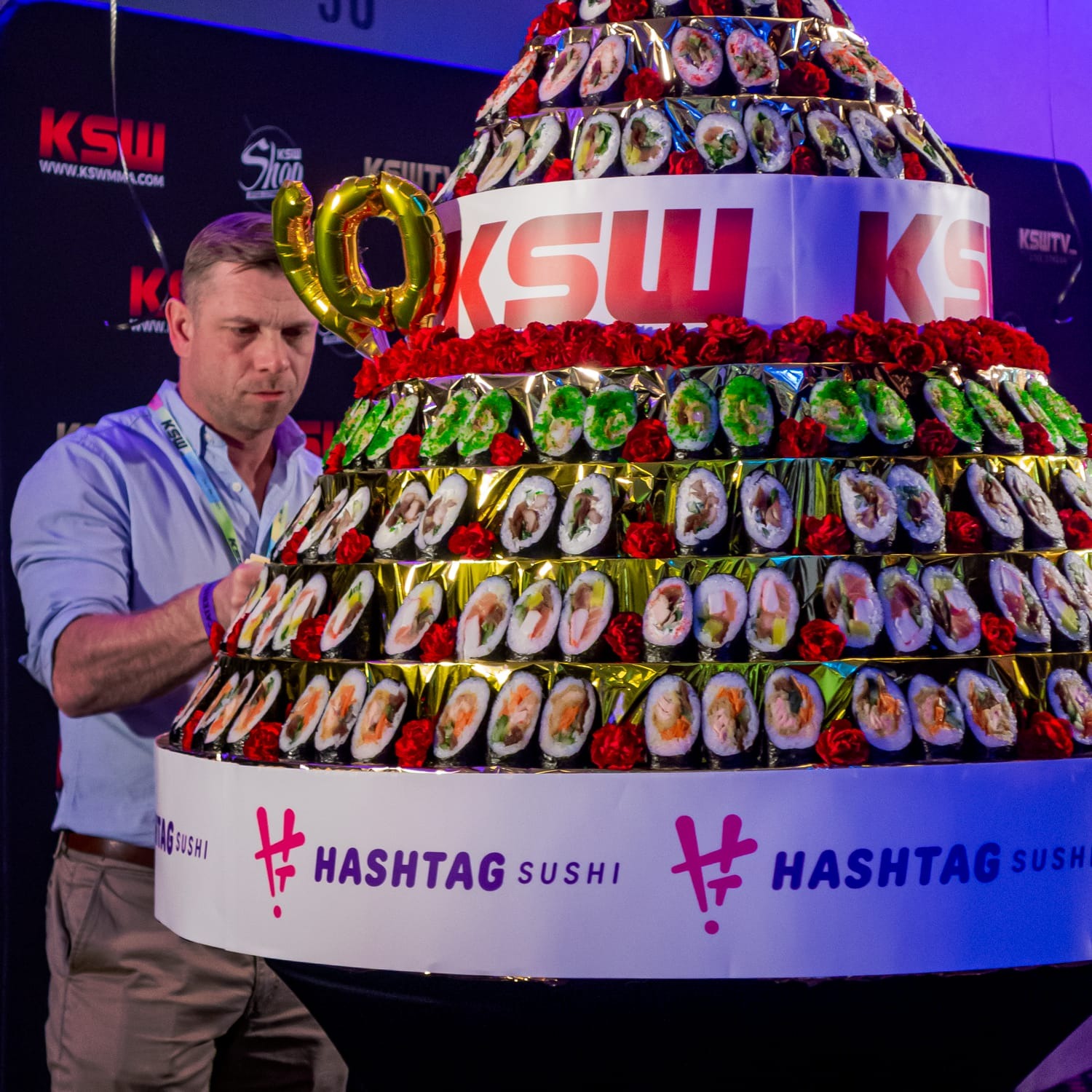 Mężczyzna w jasnej koszuli ustawia złoty balonik w kształcie cyfry '10' na piramidzie z rolek sushi udekorowanej czerwonymi różami i logotypami #hashtagsushi.