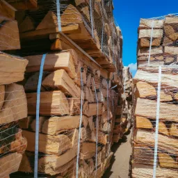 Ułożone w rzędach i zabezpieczone siatką palety z pociętym drewnem opałowym, widok z dołu na tle błękitnego nieba.