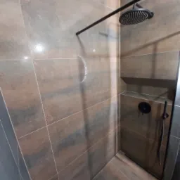 Kabina prysznicowa z brązowymi płytkami imitującymi drewno, czarna armatura, deszczownica i półka w ścianie.