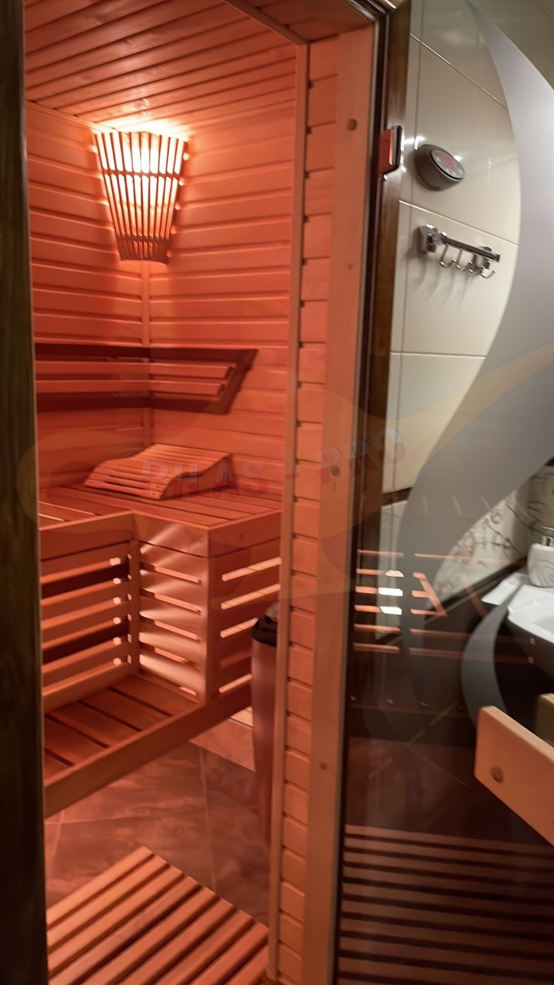 Wnętrze sauny z drewnianymi ławkami, zagłówkiem i oświetleniem, widoczne przez szklane drzwi, obok łazienka z umywalką i akcesoriami.