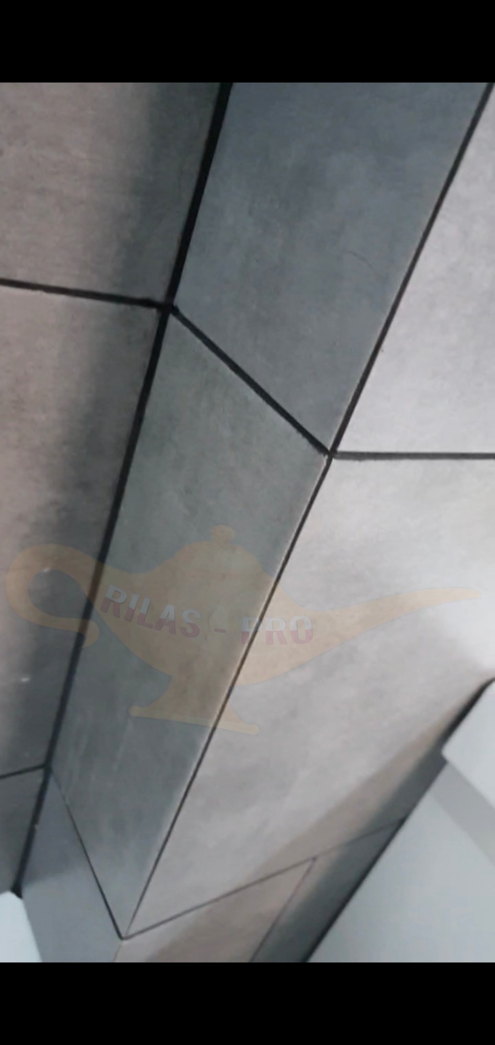 Szare płytki ceramiczne ułożone na ścianie, widoczne fugi i narożnik, z delikatnym logo firmy w tle.