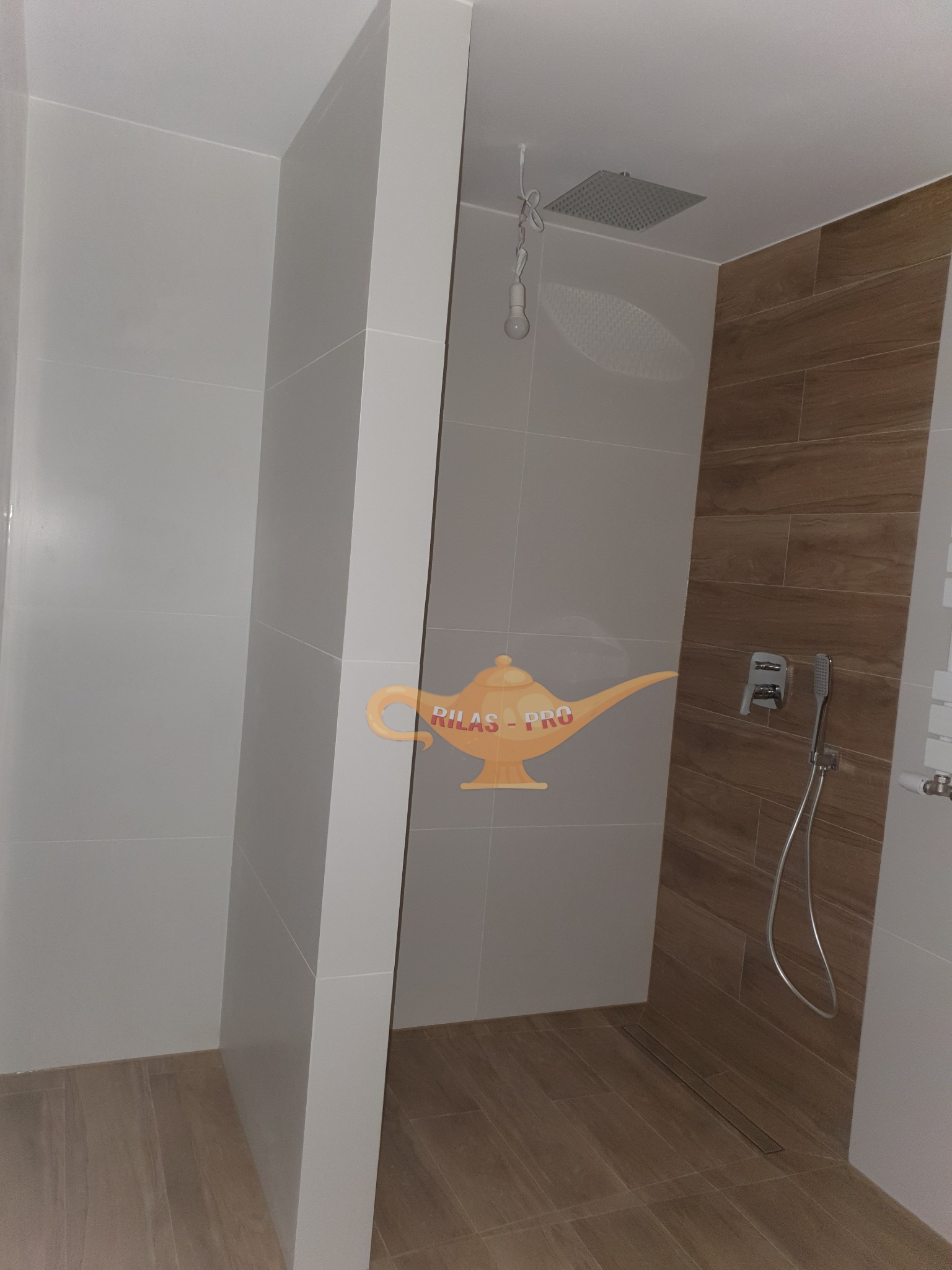 Nowoczesna kabina prysznicowa z deszczownicą i odpływem liniowym, wykończona płytkami w odcieniach szarości i drewna, z widocznym logo firmy na ścianie.