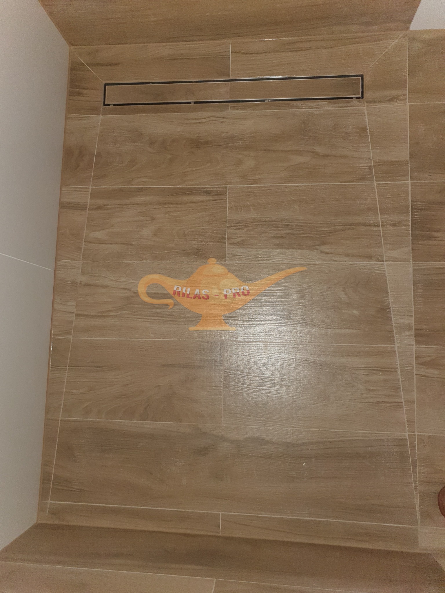 Ściana kabiny prysznicowej wyłożona płytkami imitującymi drewno z odpływem liniowym i naklejonym logo firmy w kształcie lampy Aladyna.