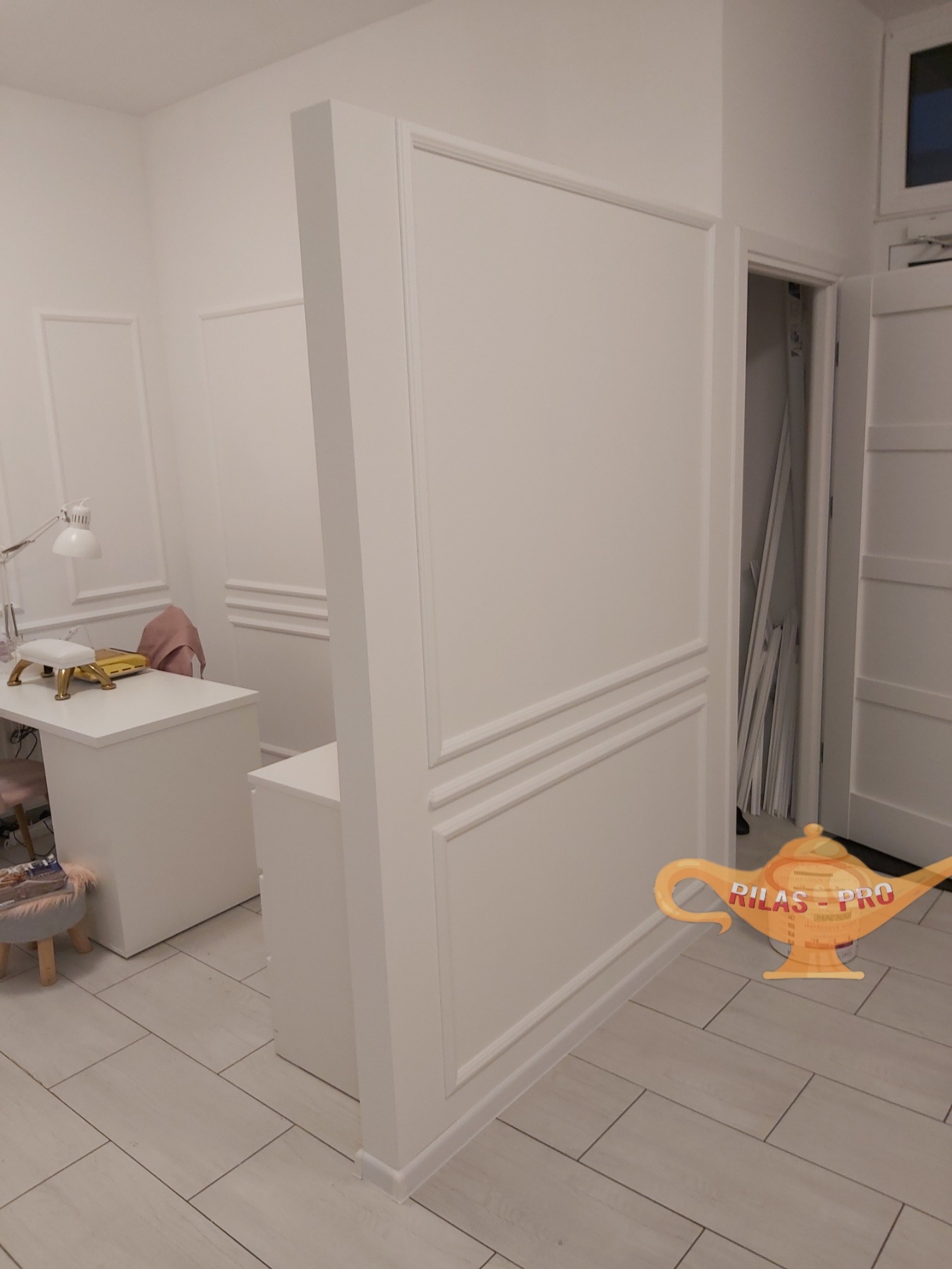 Białe panele ścienne dekoracyjne z listwami w jasnym pomieszczeniu, widoczny fragment biurka i krzesła, drzwi z białymi listwami w tle.