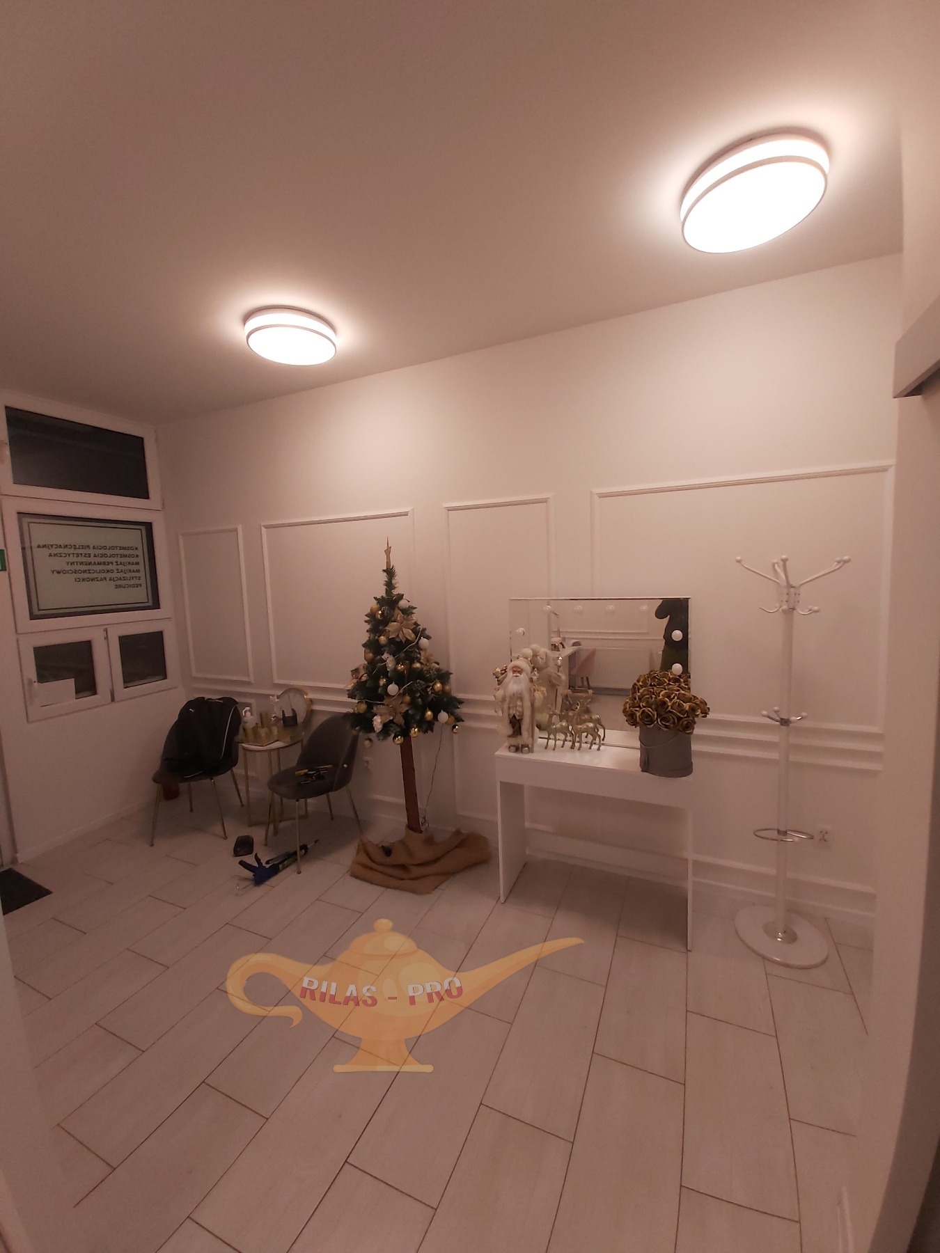 Jasne wnętrze poczekalni z choinką, białymi ścianami z dekoracyjnymi listwami, podłogą wyłożoną płytkami imitującymi drewno i minimalistycznym wyposażeniem.