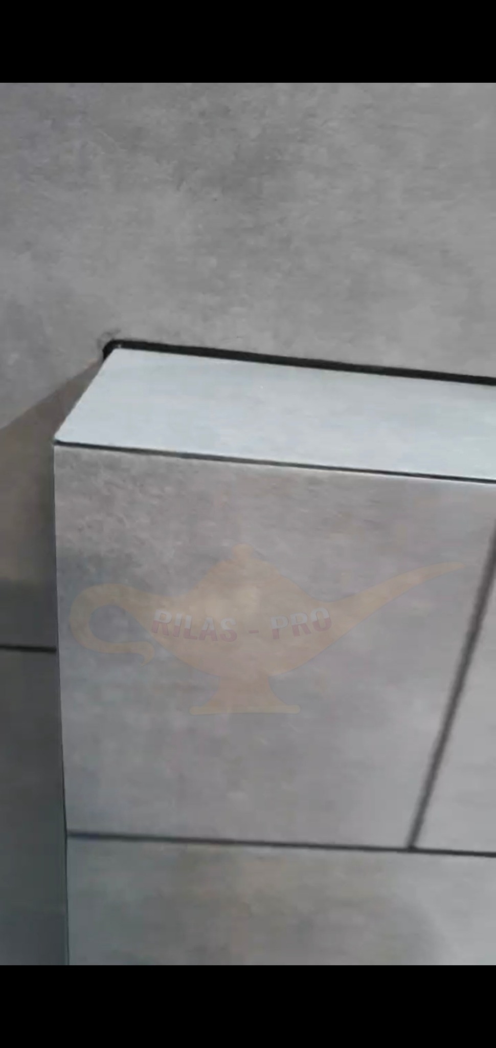 Szare płytki ceramiczne ułożone na ścianie, widoczny fragment wykończenia z czarną fugą i logo firmy nałożone na obraz.