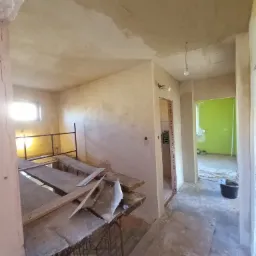 Wnętrze w trakcie prac wykończeniowych: ściany po nałożeniu gładzi, widoczne rusztowanie, narzędzia i surowa żarówka zwisająca z sufitu, w tle widoczne kolejne pomieszczenie z zieloną ścianą.