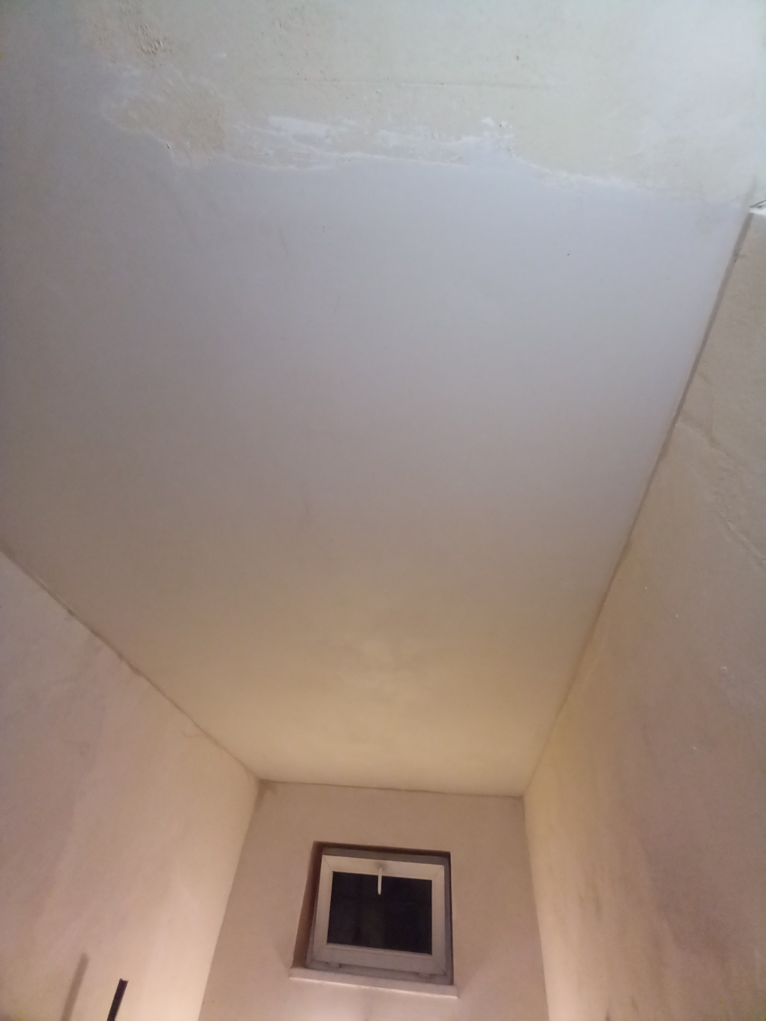 Wnętrze pokoju w trakcie remontu, widoczne ściany i sufit po szpachlowaniu, przygotowane do dalszych prac wykończeniowych, małe okno w dolnej części kadru.