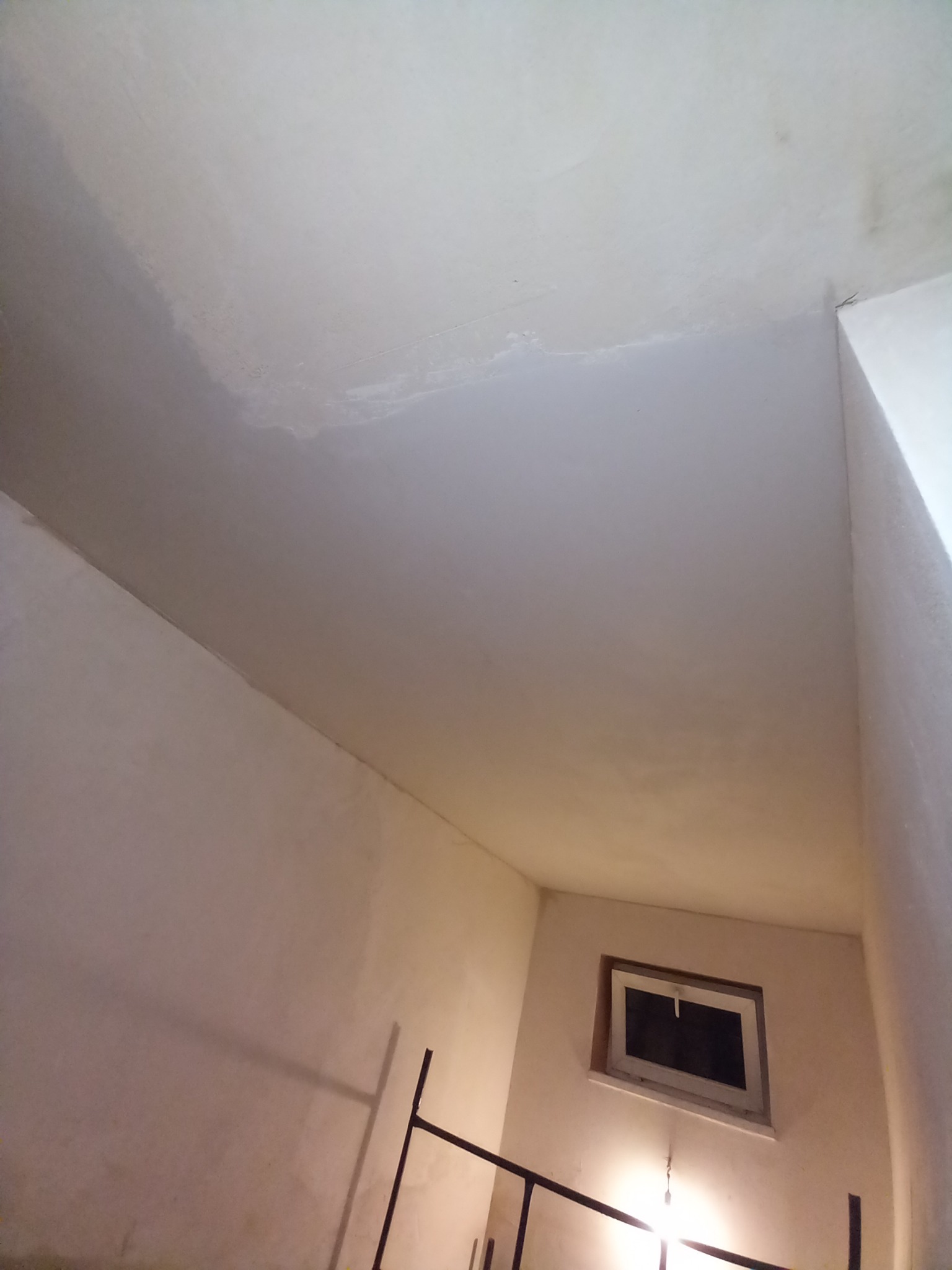 Niedokończona sufit i ściany po nałożeniu pierwszej warstwy, widoczne nierówności i ślady po szpachlowaniu, małe okno w tle.